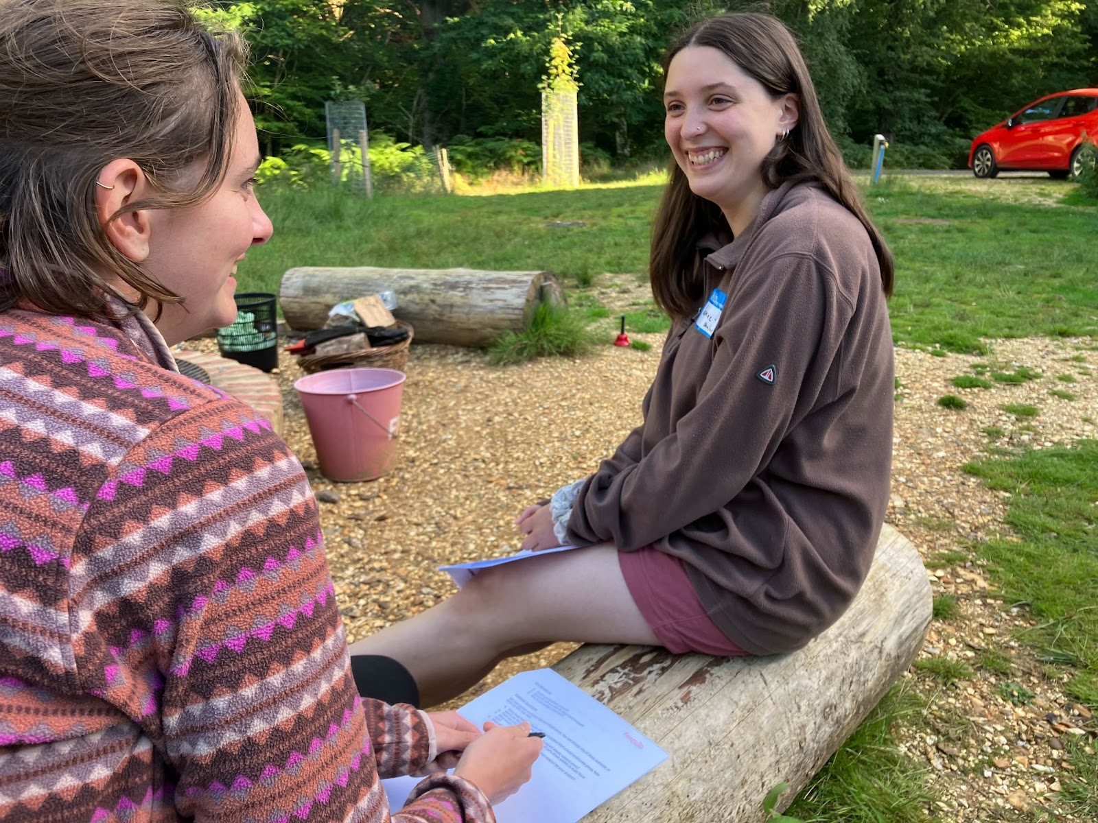 Two young women on the course sit on a log in a woodland setting, smiling and talking. They have notepaper and pens to record their thoughts.